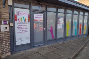 The picture is of the outside of a one storey building. There is text on the door saying "Shiloh" and pictures of multi coloured hands on the windows. 