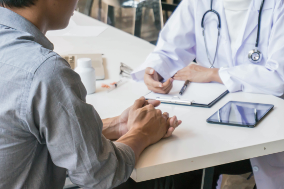 a photo of a healthcare professional sitting on a table with a patient. on the table is a clipboard, tablet and medication